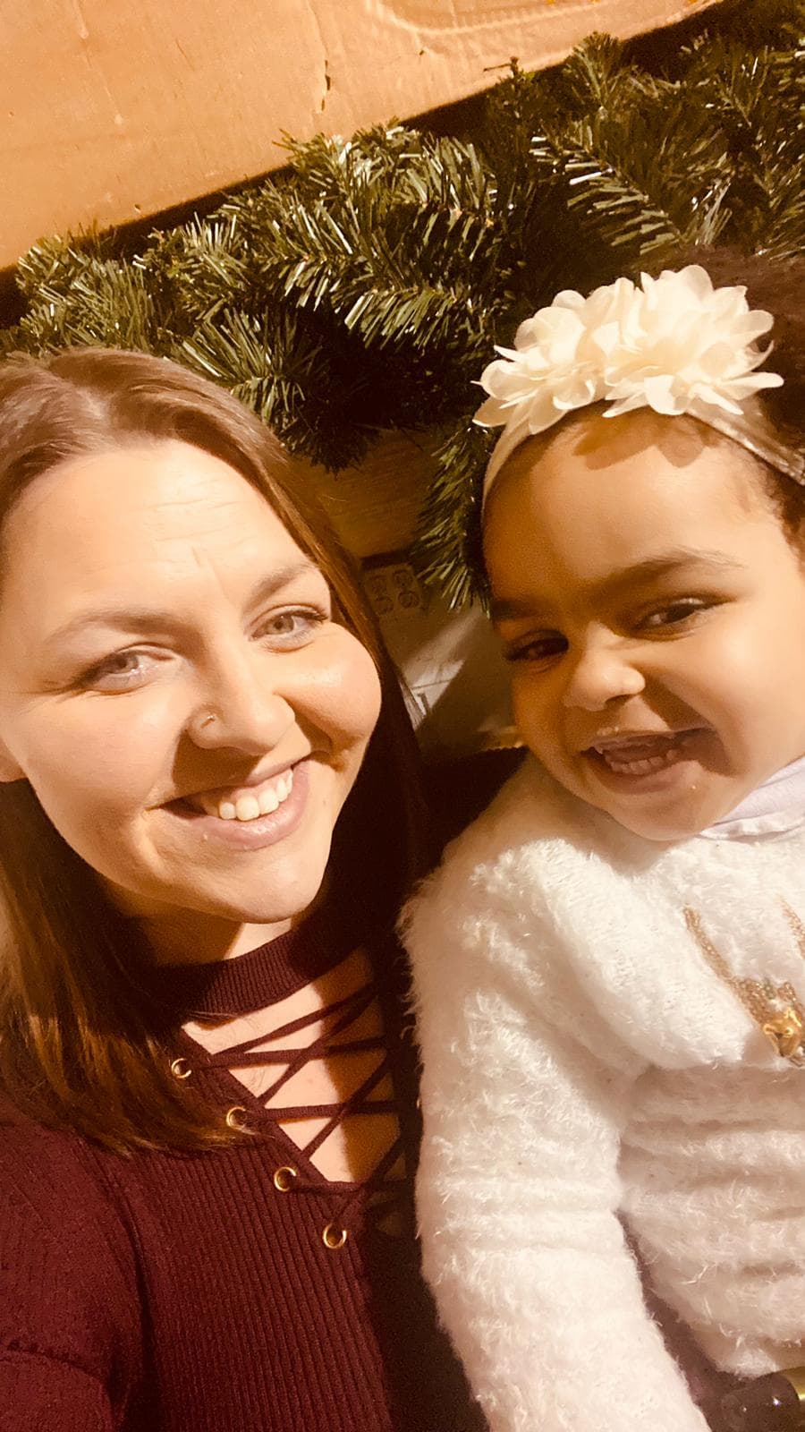 Smiling woman and toddler with a flower headband pose in front of holiday greenery.