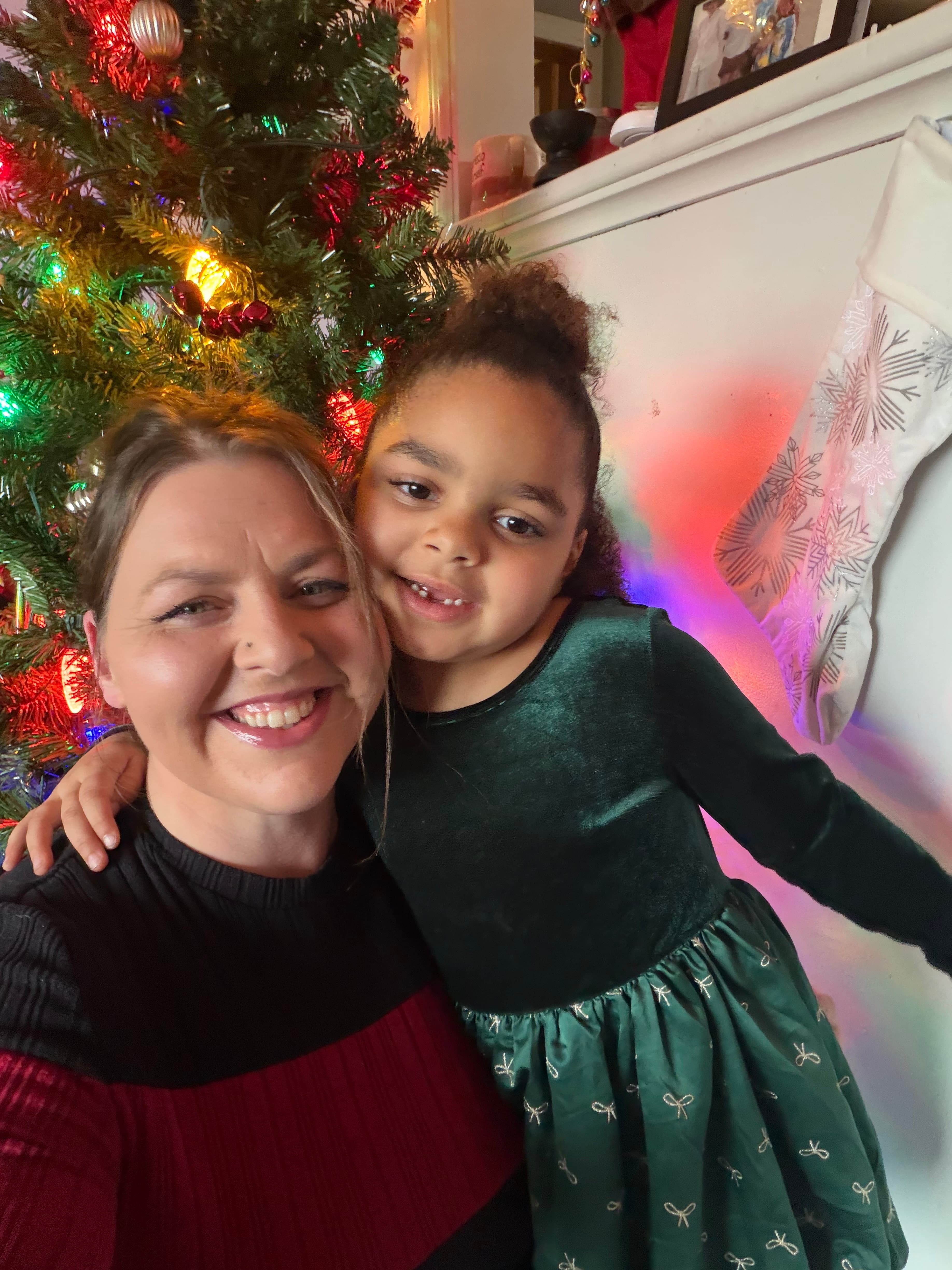 Smiling woman and young girl in festive attire pose beside a decorated Christmas tree.
